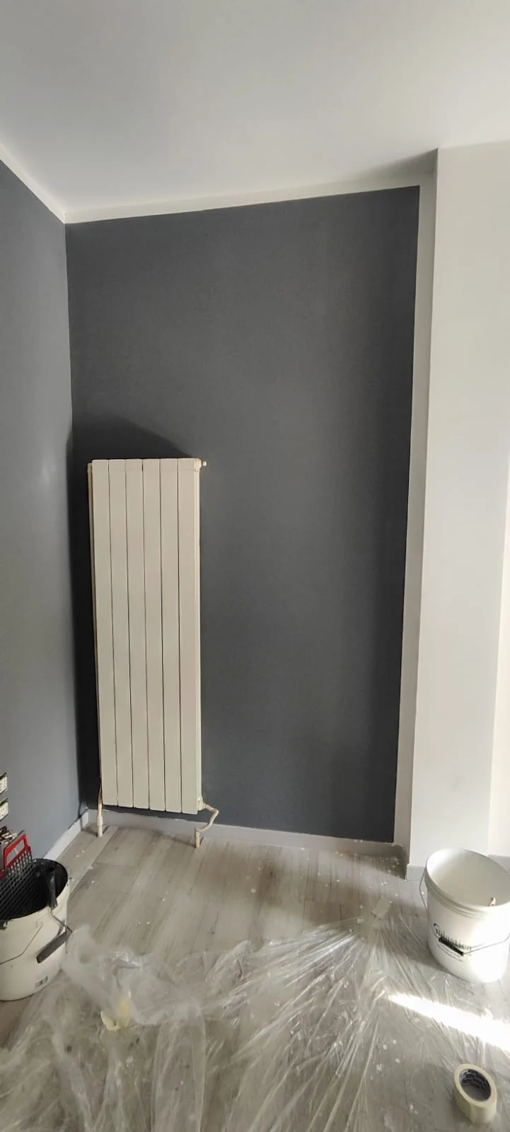 Parete grigia tinteggiata con radiatore verticale bianco e pavimento coperto dai teli